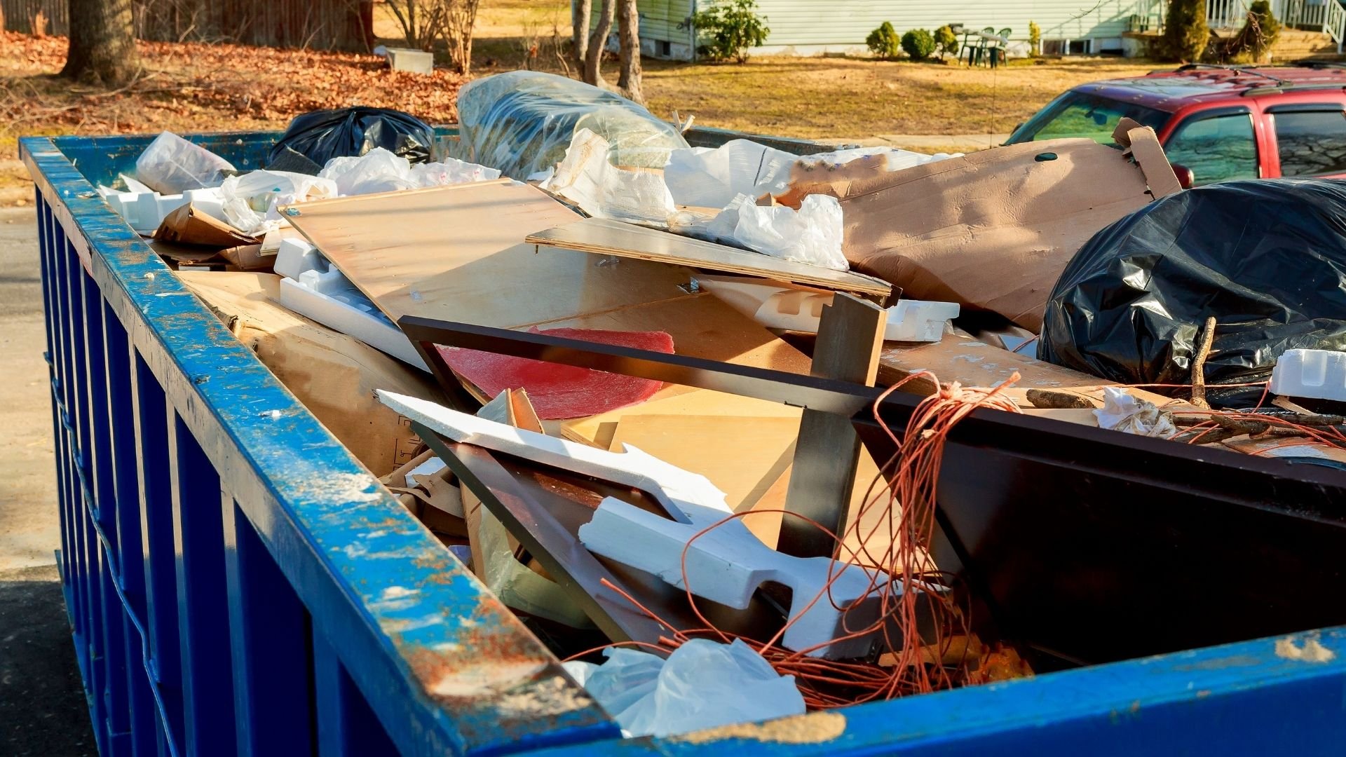 Blue dumpster filled with construction waste, wood, and debris in residential yard.