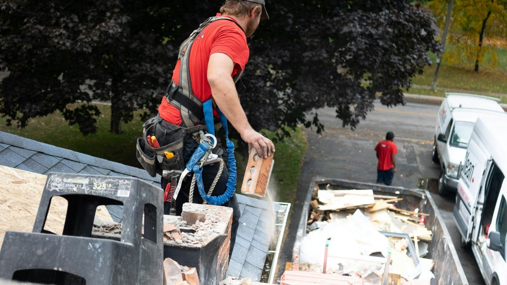 Worker in red shirt and harness removes roofing materials during demolition work.