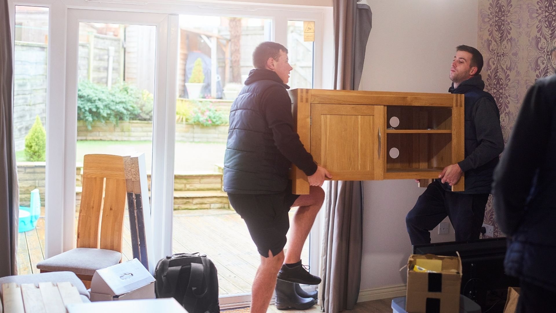 Two men moving wooden cabinet furniture indoors through glass doors.